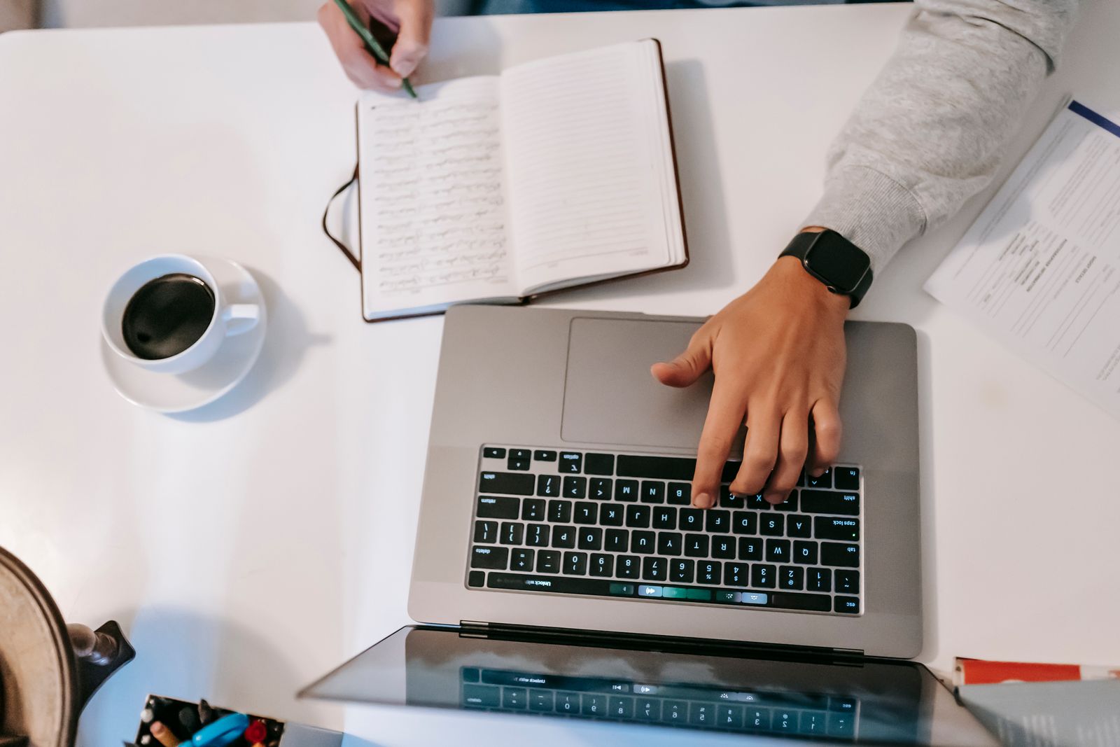 A person writing in a notebook next to an open laptop at a glass desk, planning content