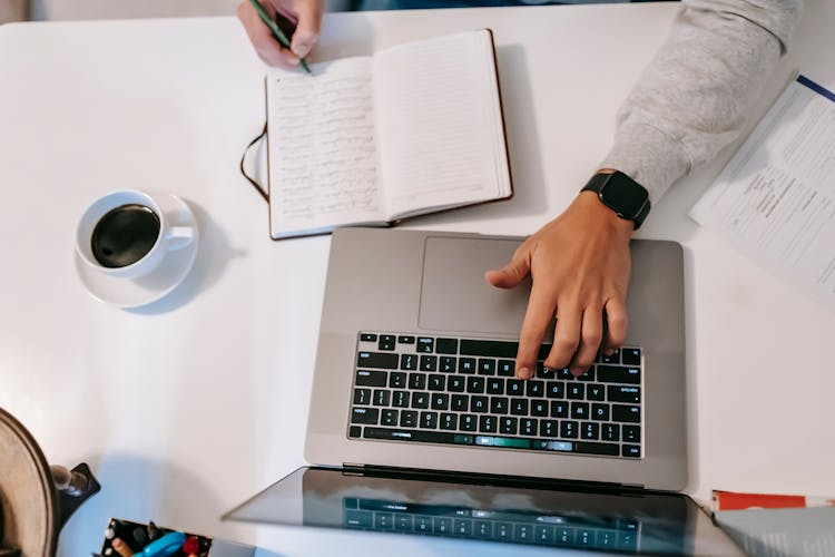 Faceless Freelancer Typing On Laptop And Writing In Journal Indoors