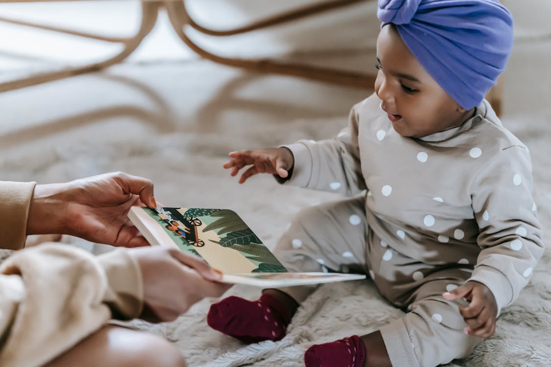 Free Side view of unrecognizable black mother showing colorful book to exited African American toddler while sitting on floor in light room on blurred background Stock Photo
