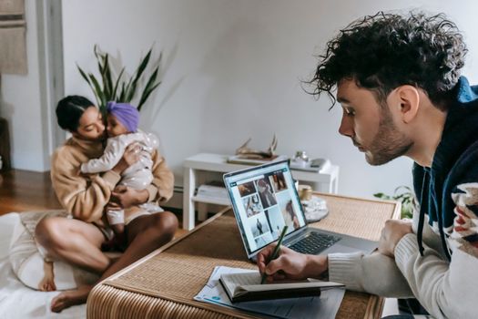 Concentrated ethnic man taking notes in notepad near modern netbook while working in room with African American woman hugging black toddler at home