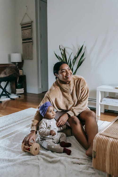 Free Cheerful African American mother and black daughter in headwear sitting on rug with toy while spending time in living room at home Stock Photo
