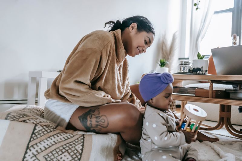 A Black mother sits beside her toddler at home and watches the child play with a wooden toy, used to illustrate reporting on autism screening and the diagnosis gap for Black children.