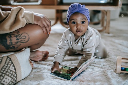 Cute African American toddler on rug with interesting book looking at camera while spending time in room with anonymous black mother