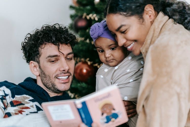 Diverse Parents Reading Book To Baby Near Christmas Tree