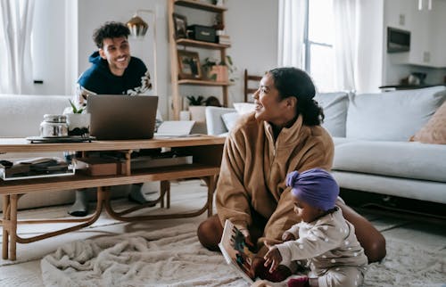 Positive African American mother and ethnic father near laptop looking at each other while spending time together at home with little black toddler