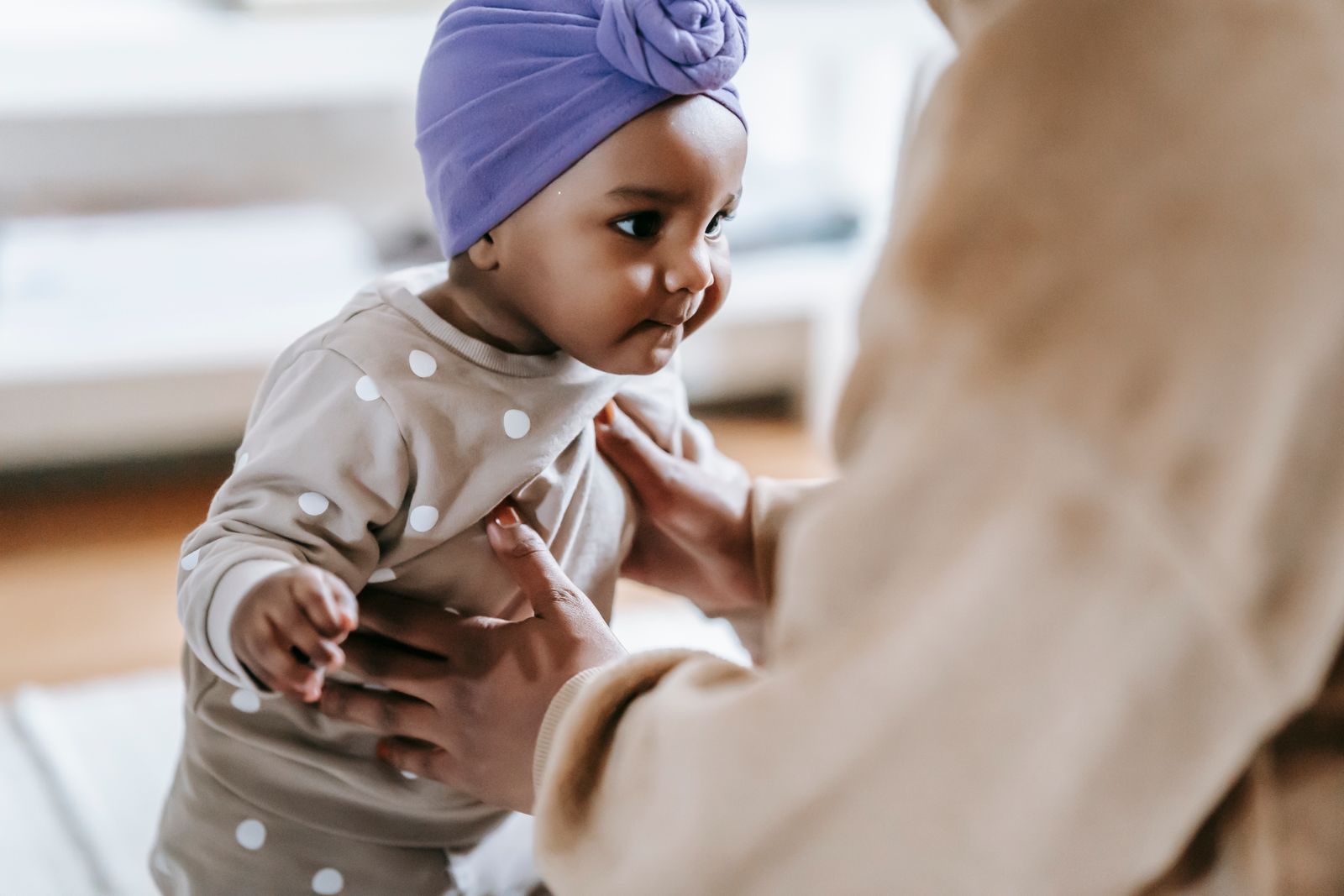 A Black mother holds her baby, who wears a polka-dot outfit and a purple head wrap, in a softly lit home. Babies under 6 months face the highest risk of severe RSV.