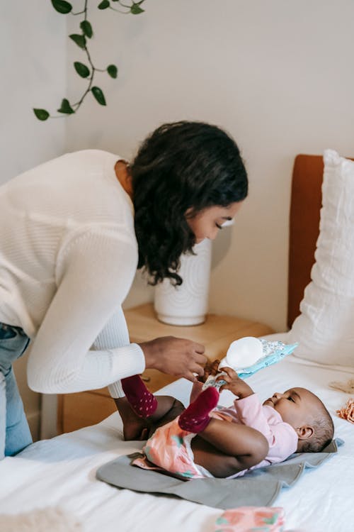 Cheerful ethnic mother touching hands of small adorable African American baby lying on bed