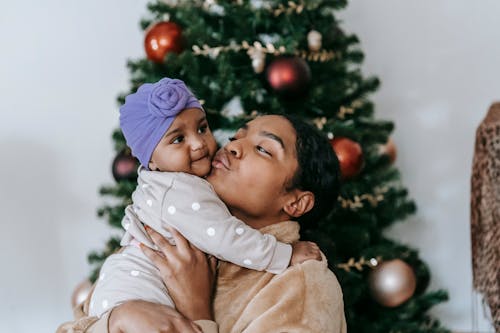 Side view of African American mum pouting lips for kissing small girl in turban against decorated Christmas tree