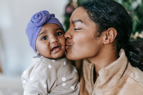 Free Gentle African American mum with closed eyes kissing adorable little daughter in turban at home Stock Photo
