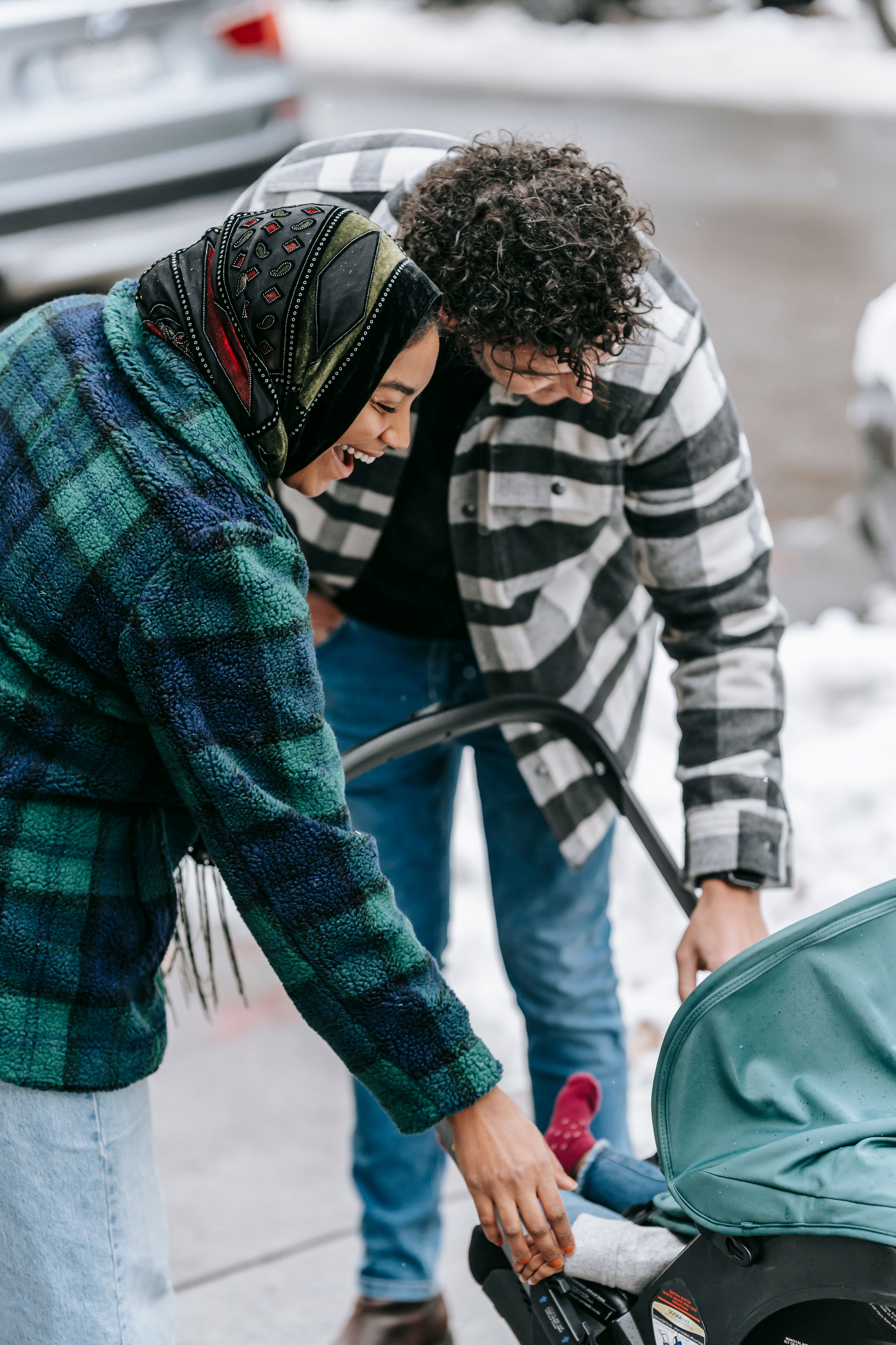 Happy ethnic couple in casual clothes standing near stroller and looking at baby on street in daytime