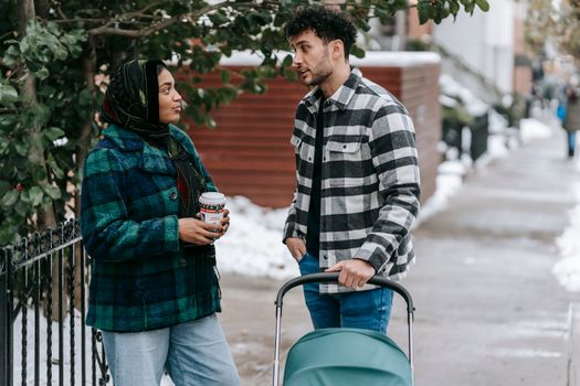 Ethnic woman wearing headscarf standing with cup of takeaway drink and talking to husband standing near stroller on snowy street