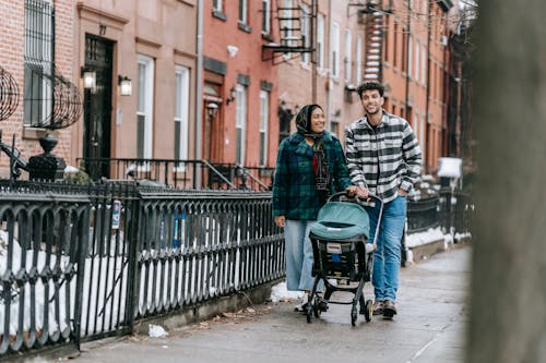 Full body of positive ethnic couple pushing stroller with baby while strolling along street in winter day