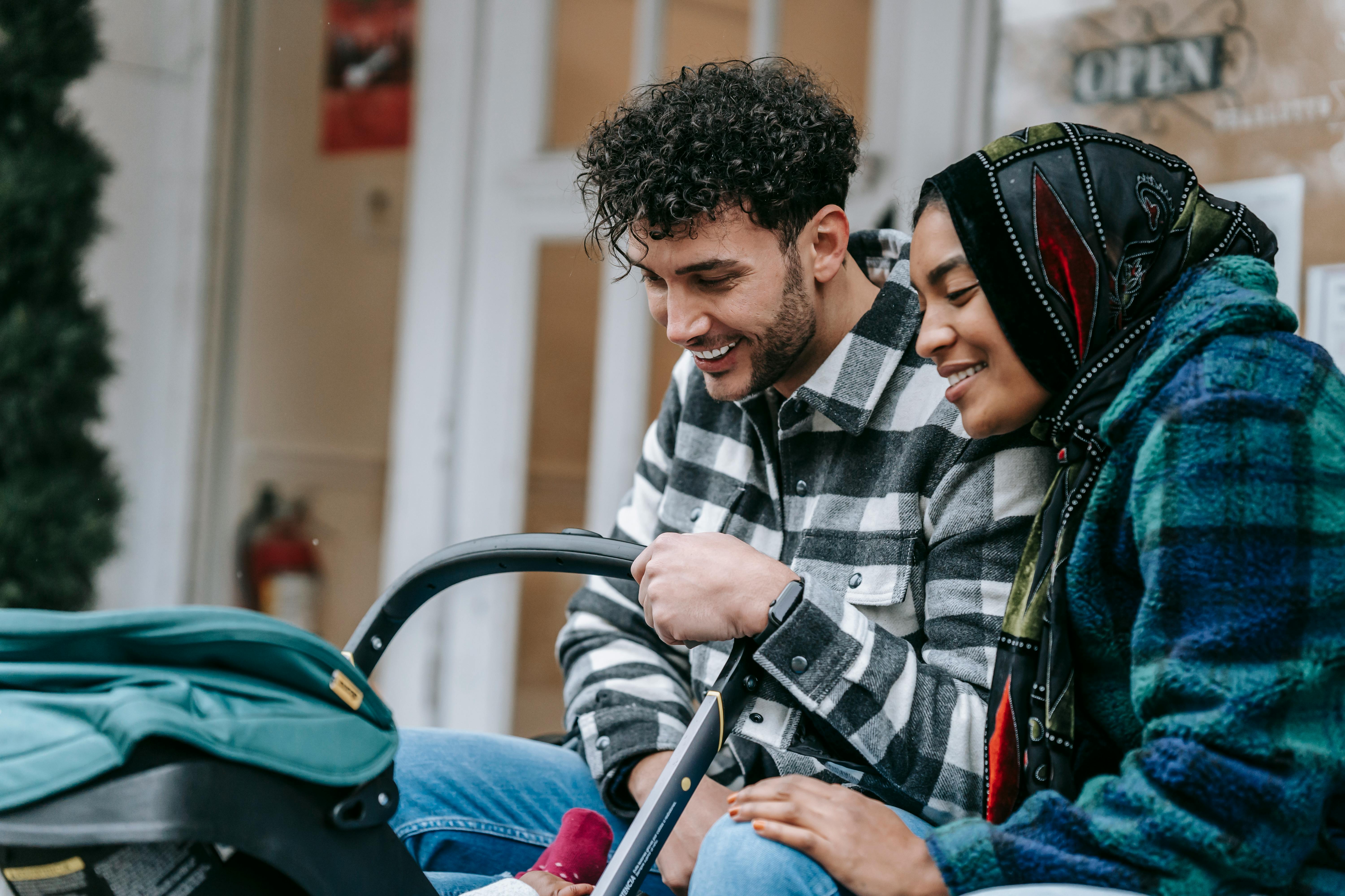 cheerful young multiethnic spouses smiling and looking at baby in stroller