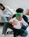 Unrecognizable young father sitting n floor and helping cute little black daughter to walk near smiling ethnic mother sitting on sofa