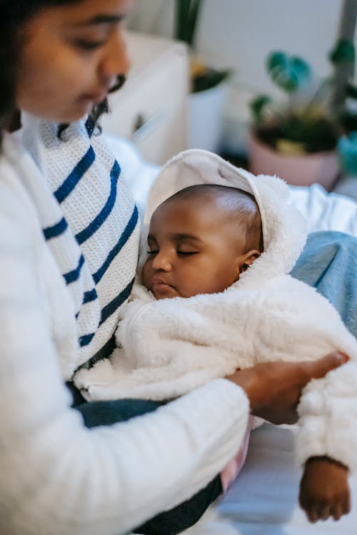 Free Side view of crop young African American female in casual clothes hugging cute little sleeping baby while sitting on sofa at home Stock Photo
