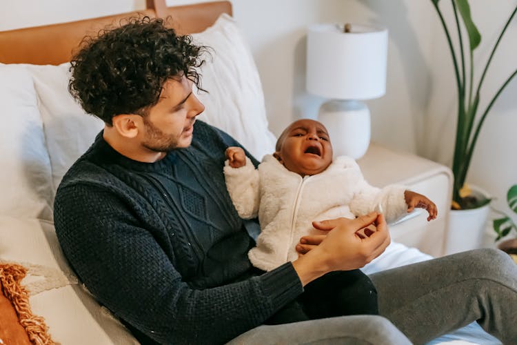 Young Ethnic Man Sitting On Bed With Crying Baby In Arms