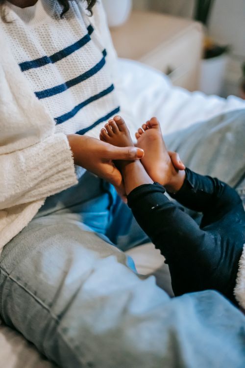 Free Crop unrecognizable African American female in casual clothes sitting on comfortable bed and caressing cute barefooted toddler Stock Photo