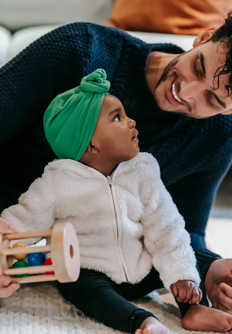 Smiling Young Ethnic Man Playing With Curious Baby On Carpet At Home