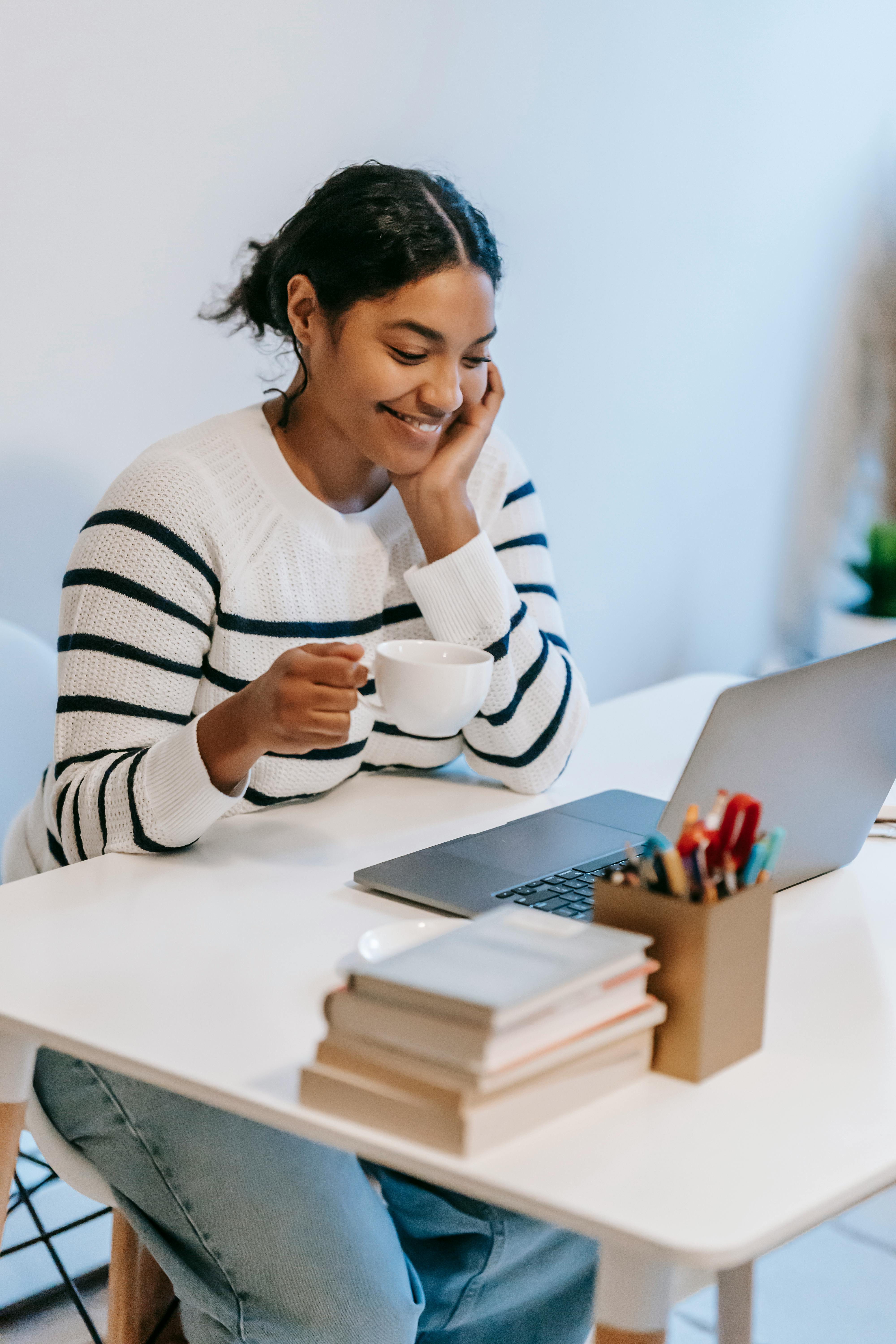 a girl with a cup, books, stationery, and a mobile phone, looking at her laptop.