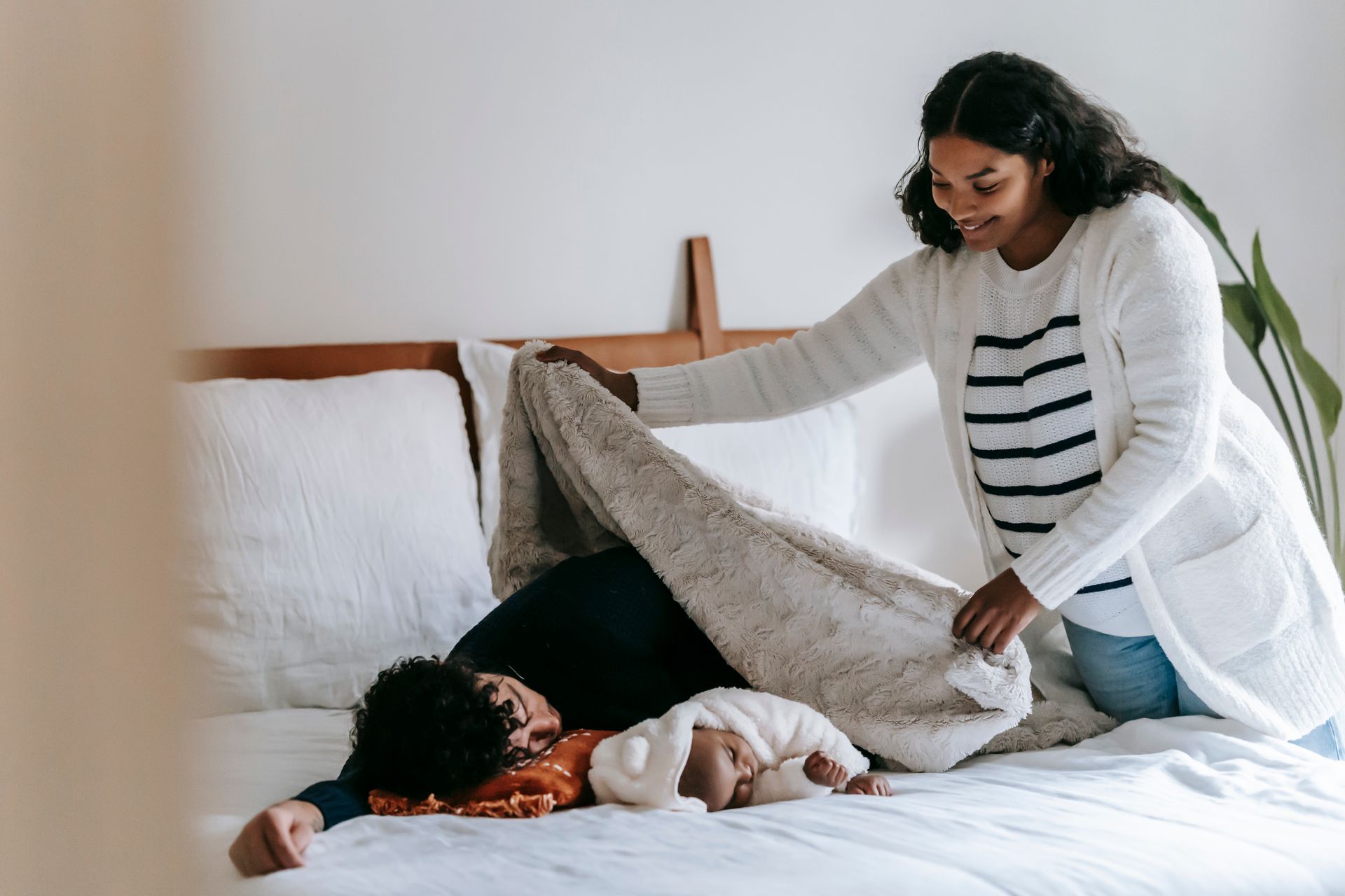 Mother covers her baby and partner with a blanket in a peaceful bedroom setting, fostering familial warmth and affection.
