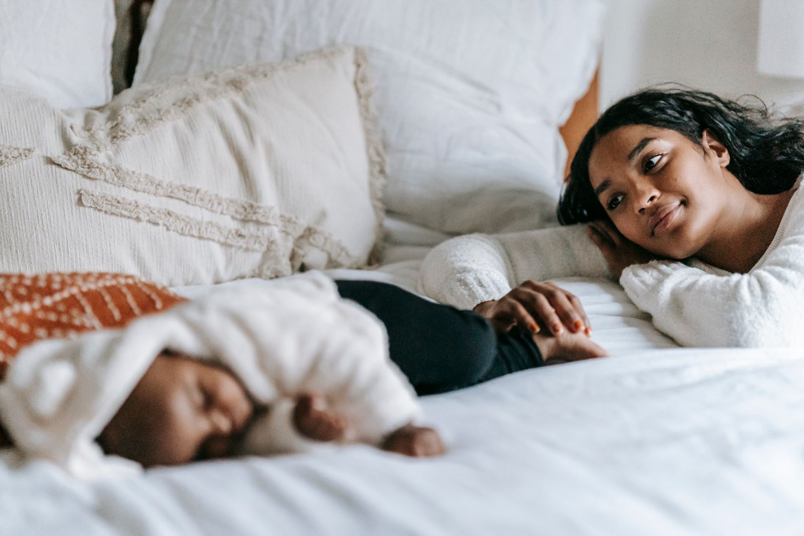 A Black mother lies on a cream-colored bed beside her sleeping Black infant, gazing at the baby with a peaceful contemplative expression in a sunlit domestic interior.