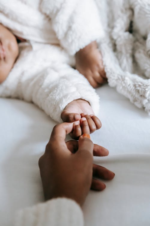 High angle of crop unrecognizable African American woman holding hand of newborn baby baby sleeping on soft bed in daylight