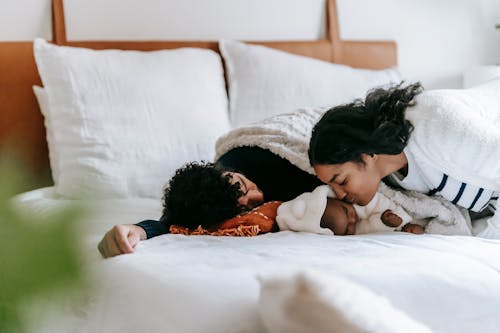 Free Happy young African American mother kissing adorable baby sleeping on cozy bed with ethnic father in sunny morning Stock Photo