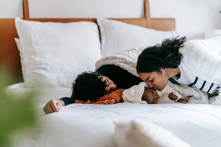 Young Black Woman Kissing Baby Sleeping On Bed Near Father