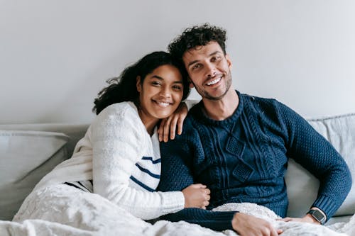 Happy ethnic couple in casual clothes sitting on soft sofa in light room and looking at camera in daytime
