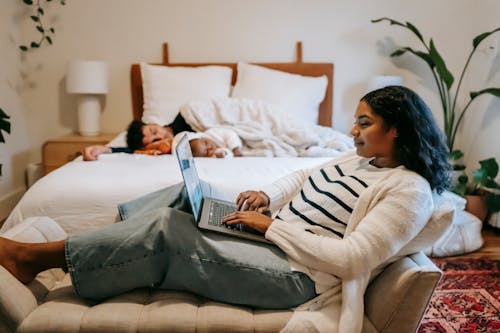 Free Side view of pensive ethnic woman freelancer sitting on couch and browsing netbook while man and little baby sleeping on bed in cozy bedroom Stock Photo