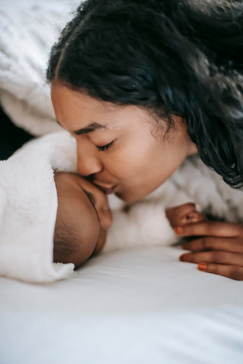 Calm ethnic mother kissing little baby lying on bed and sleeping at home in daylight