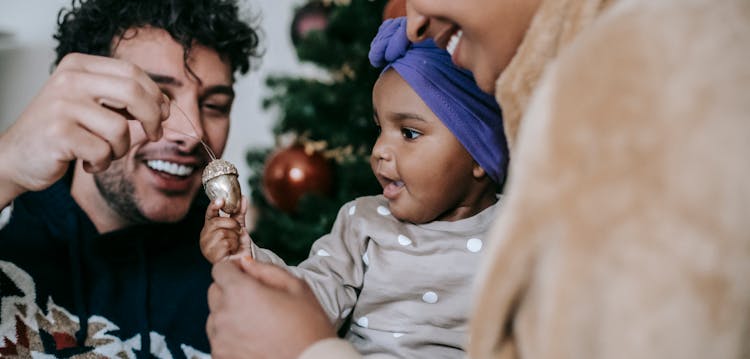 Happy Ethnic Couple Playing With Baby Against Christmas Tree At Home