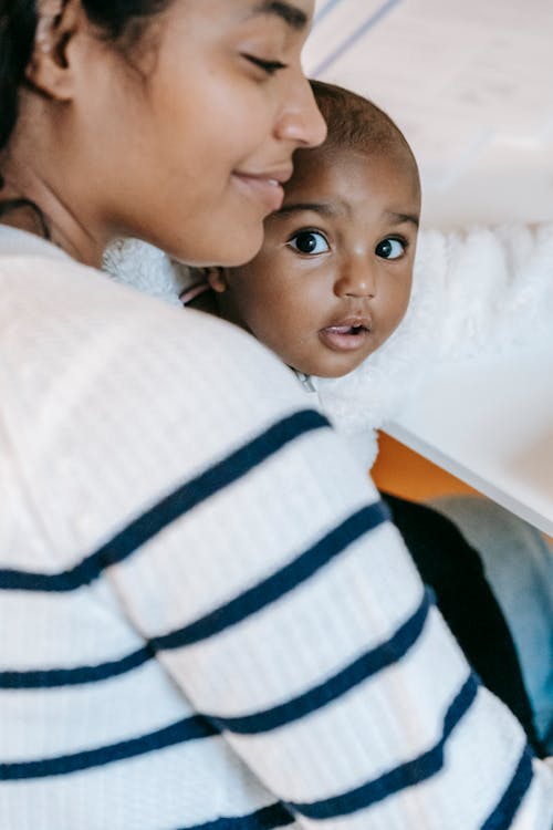Free High angle side view of crop smiling gentle African American mom cuddling curious baby with open mouth Stock Photo