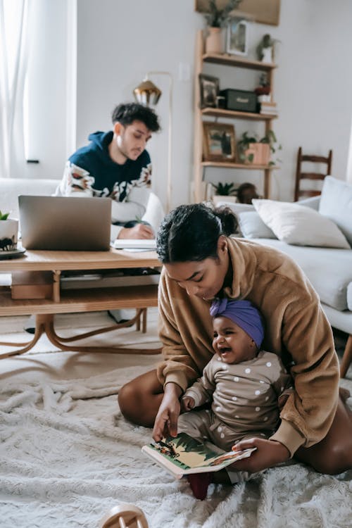 African American mother and black baby sitting on rug with book in living room near ethnic father at table with laptop