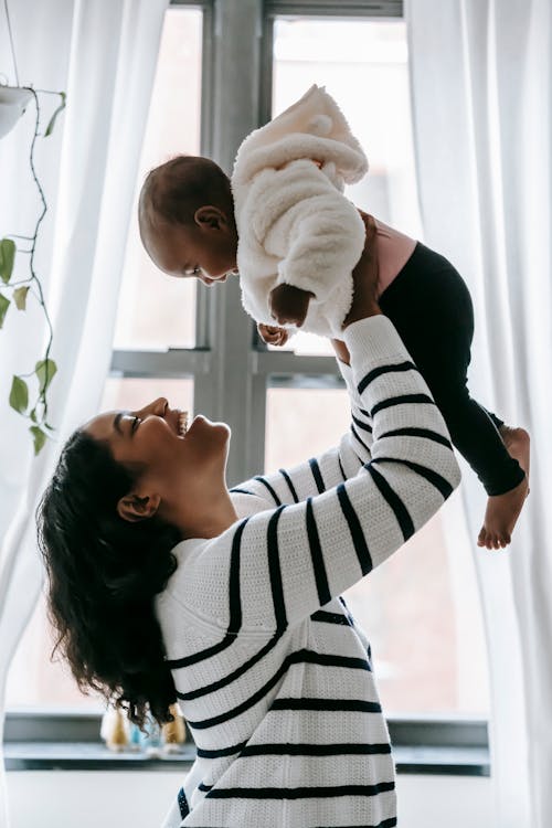 Side view of cheerful African American mother and cute black baby in hands looking at each other while standing near window