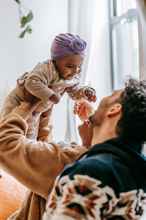 Side view of anonymous multiethnic parents with cheerful little African American baby in hands spending time together in living room