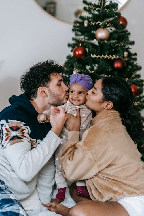 Tender multiracial parents caressing little black baby while sitting near decorated fir tree in room during Christmas celebration at home