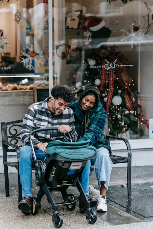 Full body of smiling multiracial parents sitting on bench with anonymous child in baby stroller on street near showcase in city