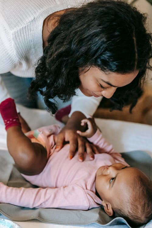 From above of tender African American mother putting clothes on cute black baby lying on bed in bedroom on blurred background