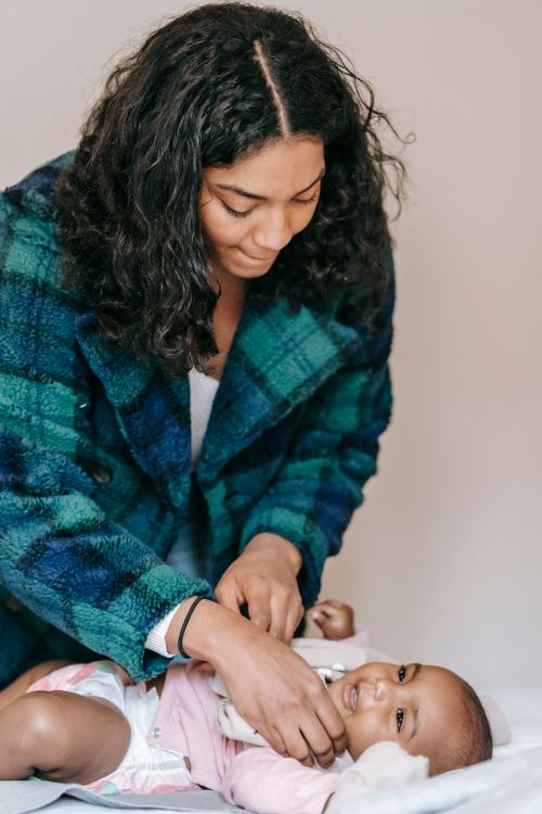 Free Focused African American mother putting clothes on cute little black positive baby lying on bed in light bedroom at home Stock Photo