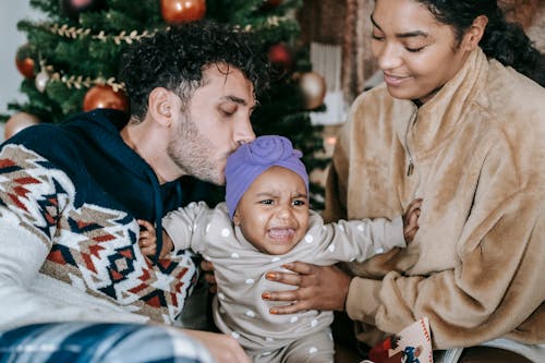 Free Tender multiracial parents kissing little unhappy baby with headband while sitting near decorated spruce in room during Christmas celebration at home Stock Photo