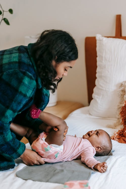 Side view of serious African American mother putting clothes on unhappy black baby lying on bed in bedroom at home