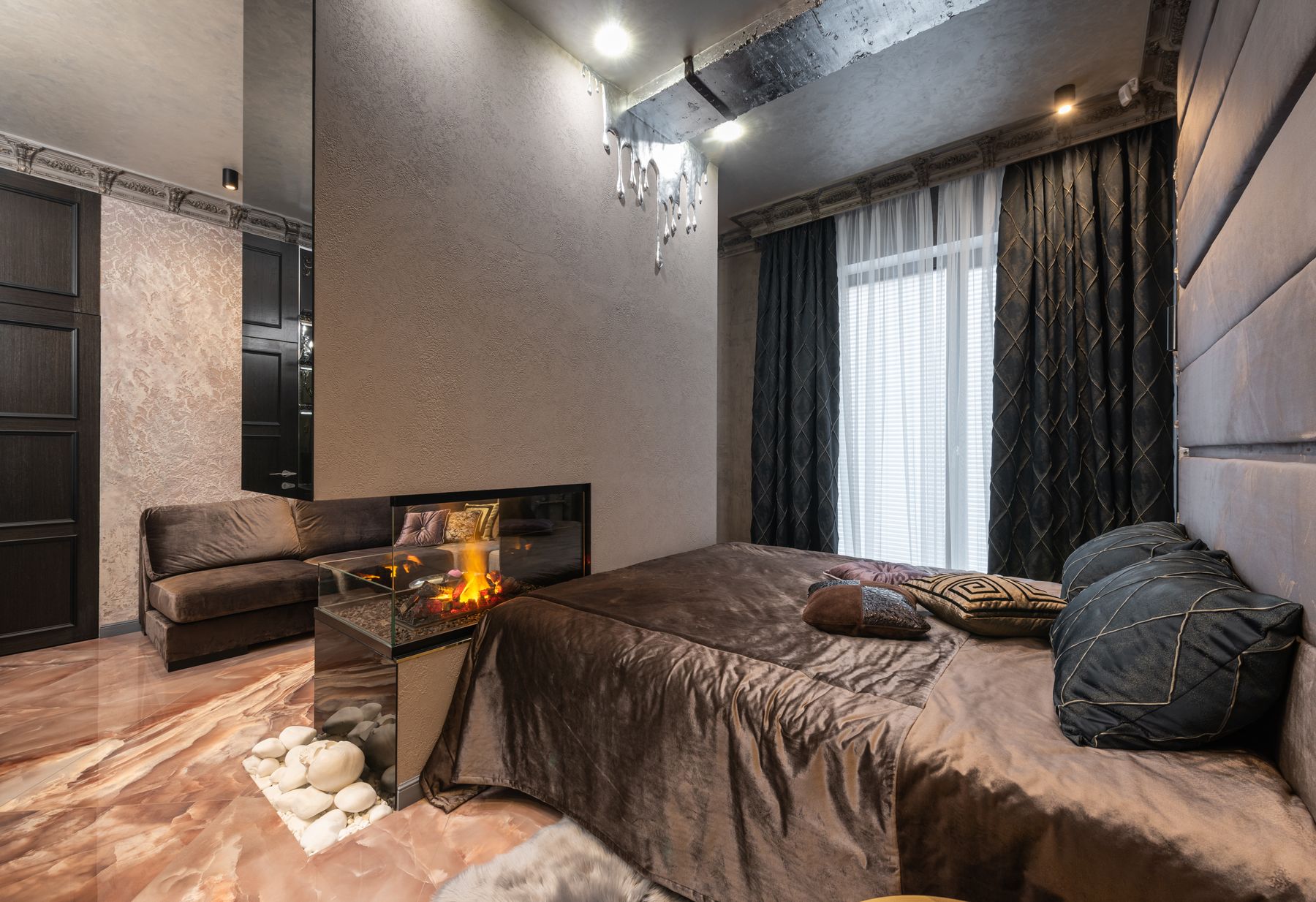 Spacious bedroom with a pale bed, sofa, brown textiles and a lit fireplace on the adjoining wall