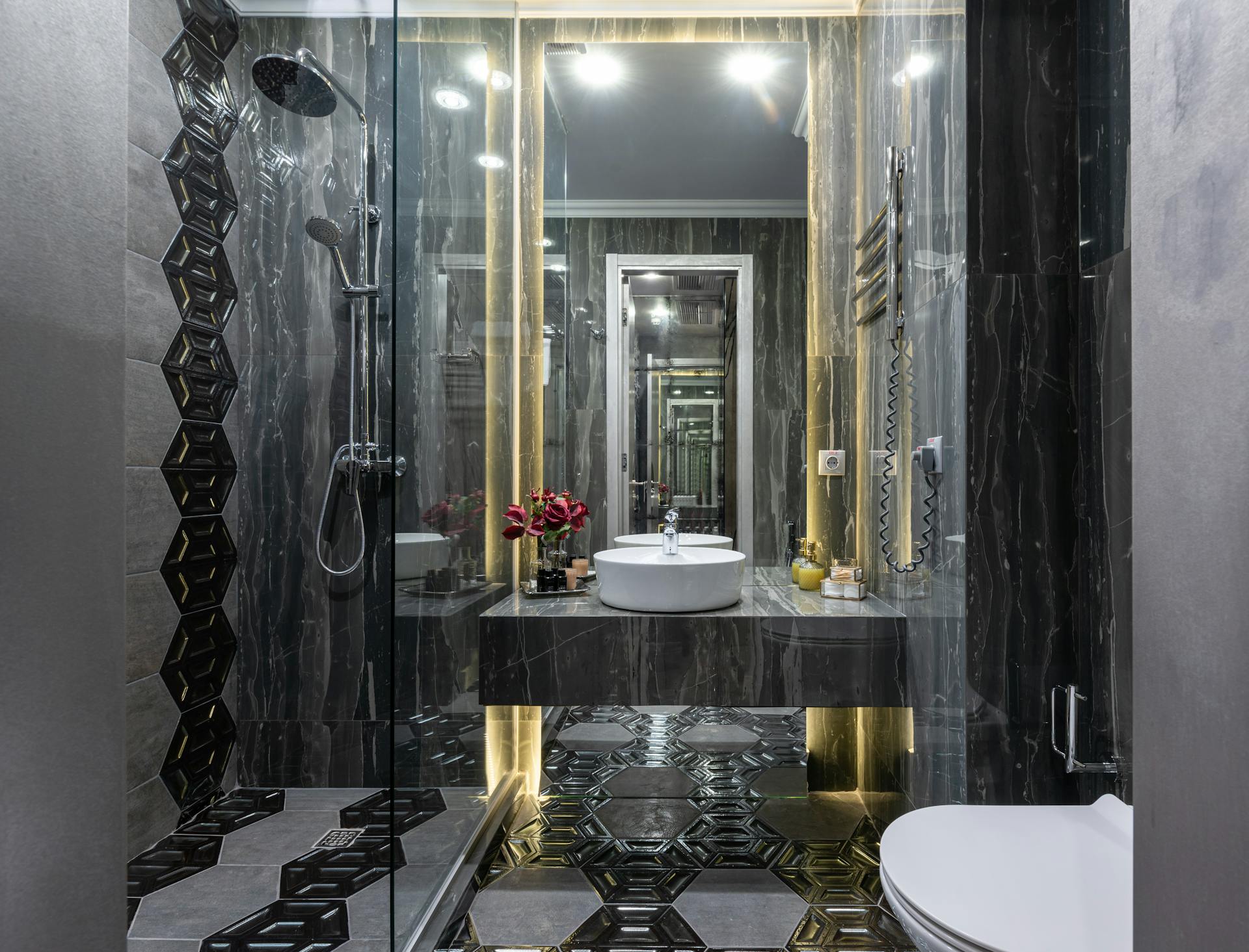 Interior of spacious contemporary bathroom with black tiled walls and floor with transparent shower cabin near bidet and sink at mirror