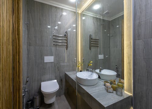 Interior of stylish restroom with gray walls and ceramic bidet near white sink reflecting in big mirror in light bathroom