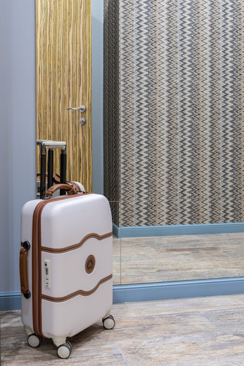 Modern baggage placed on floor near reflective mirror against wall with wooden door in corridor of hotel room during trip