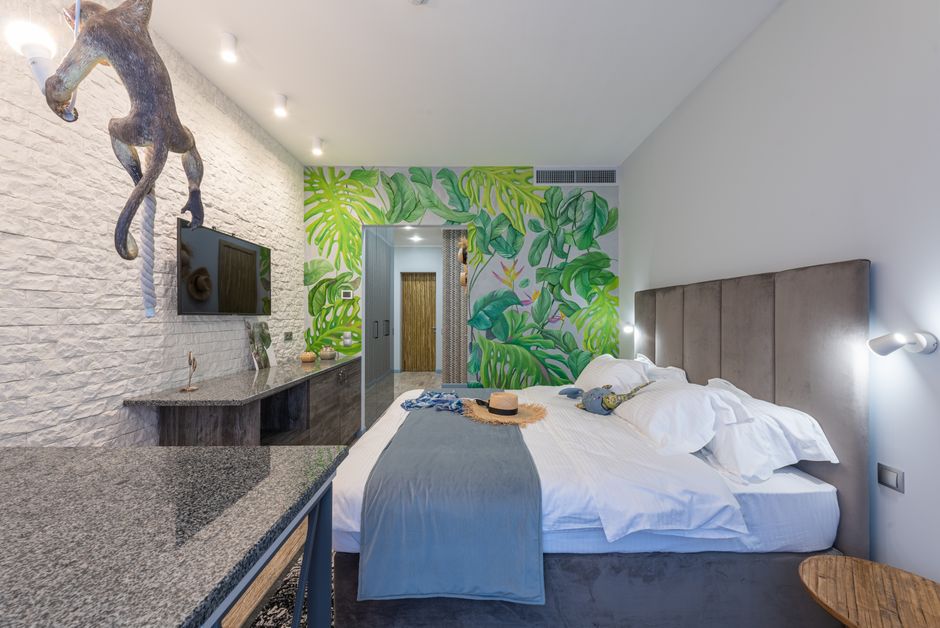 Straw hat on comfortable bed with blanket in spacious hotel room with TV hanging on decorated wall and green painted wall