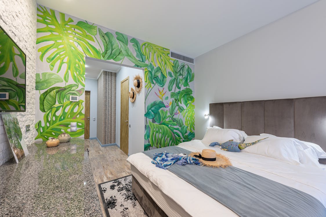 Free Comfortable Bed With Straw Hat Placed In Hotel Suite With Decorations And Green Painted Wall During Trip In Tropical Country Stock Photo