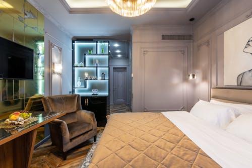 Table and armchair against bed with cover under glowing chandelier in house bedroom with entrance door