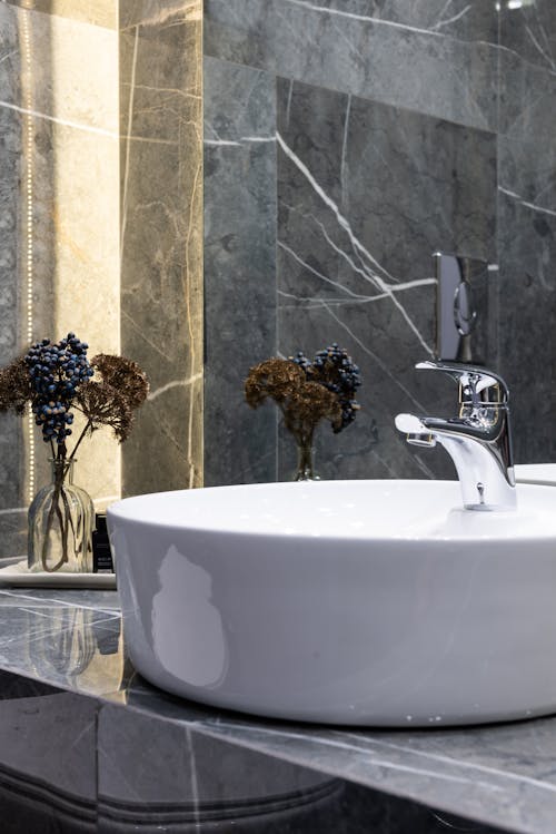 Washstand with shiny metal tap against plant sprigs in vase reflecting in mirror in modern bathroom